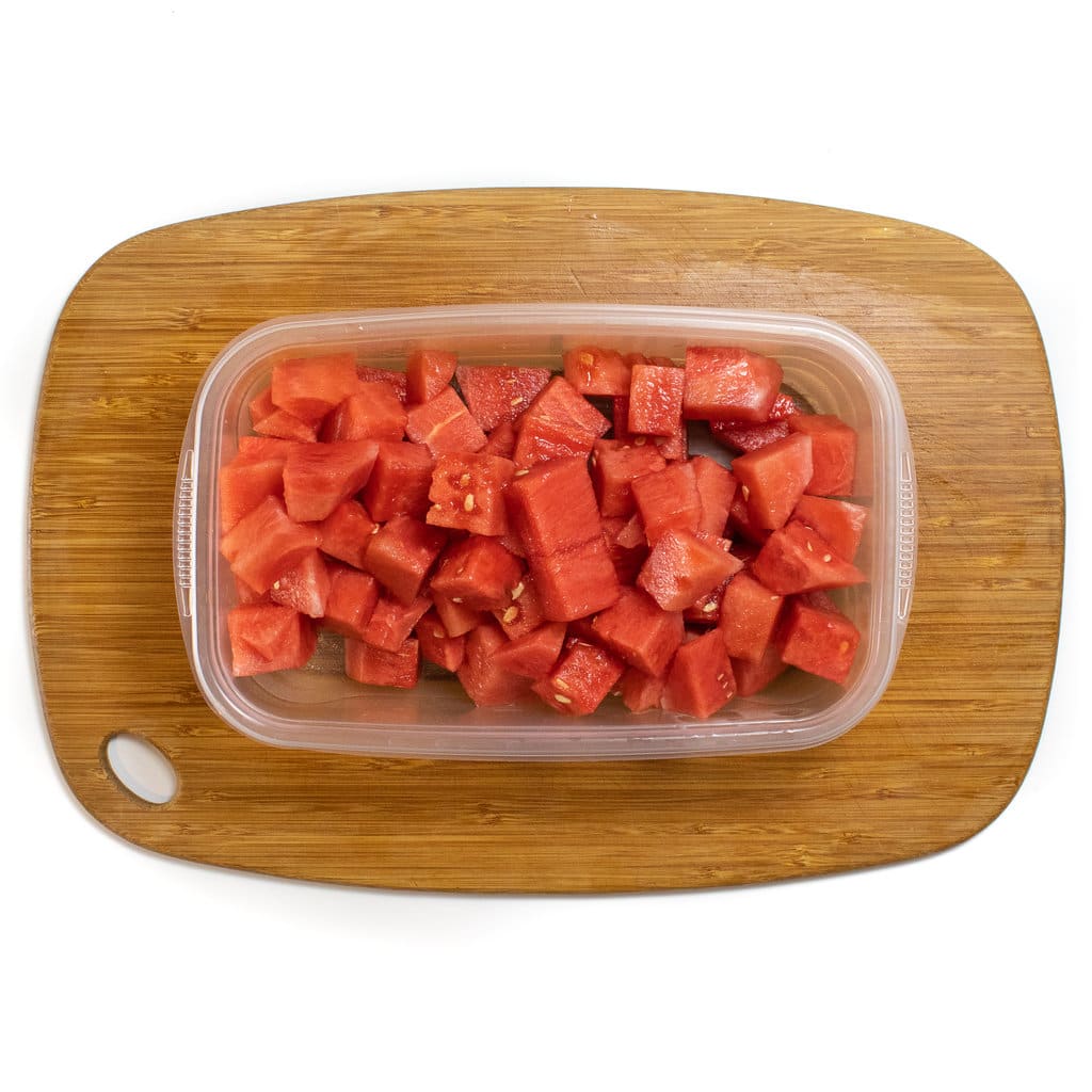 A wooden cutting board with a container full of cubes of watermelon.