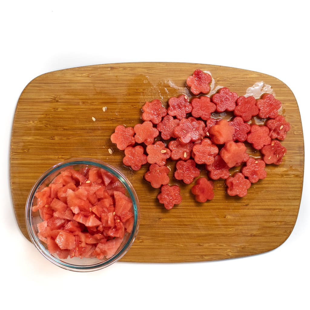 A wooden cutting board against a white background with a ton of flower shaped watermelon and a bowl of watermelon scraps.