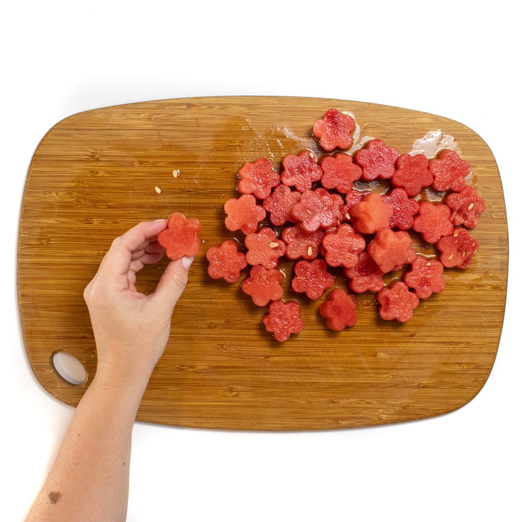 A hand holding a flower shaped watermelon piece against a wooden cutting board with a pile of flowers shaped watermelon pieces
