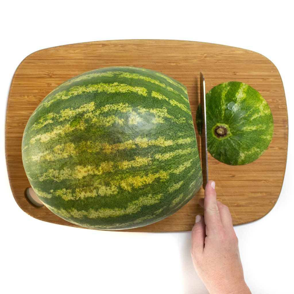 A watermelon on a cutting board with a hand slicing off the end.
