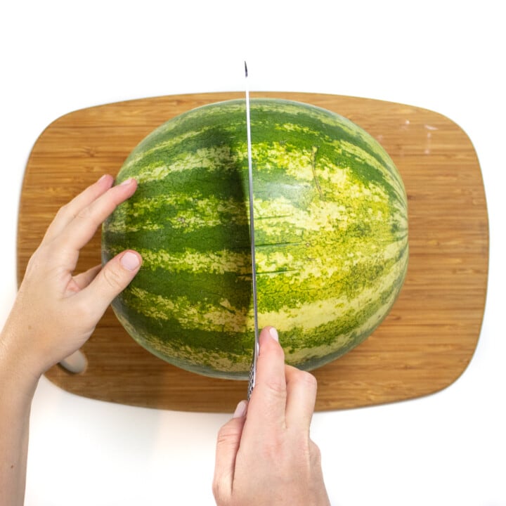 A watermelon on a wooden cutting board with hands holding a knife cutting it in half.