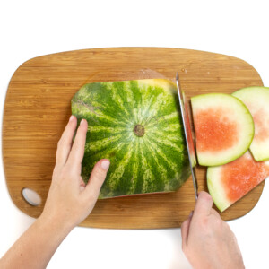 A watermelon board cut in half, with hands holding a knife cutting the edges off