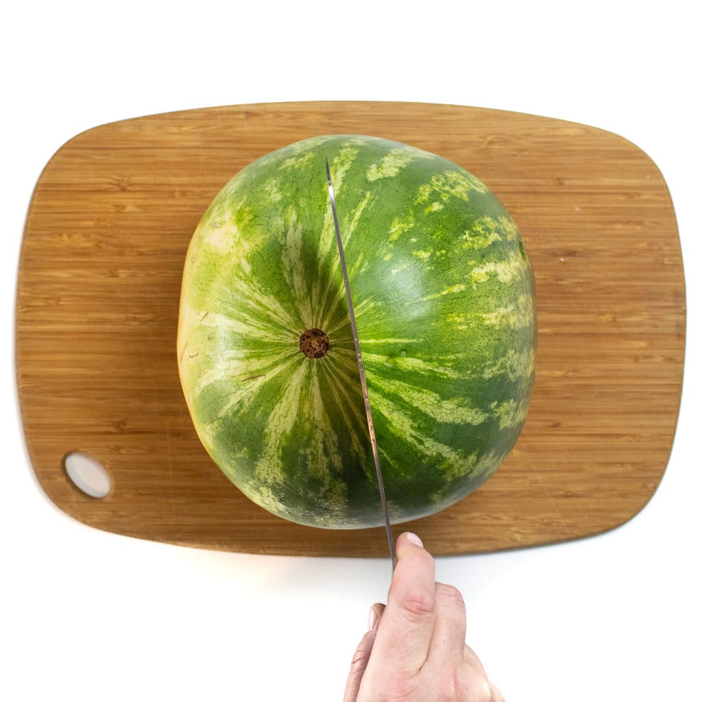 A watermelon sitting on a cutting board with a knife cutting it in half.