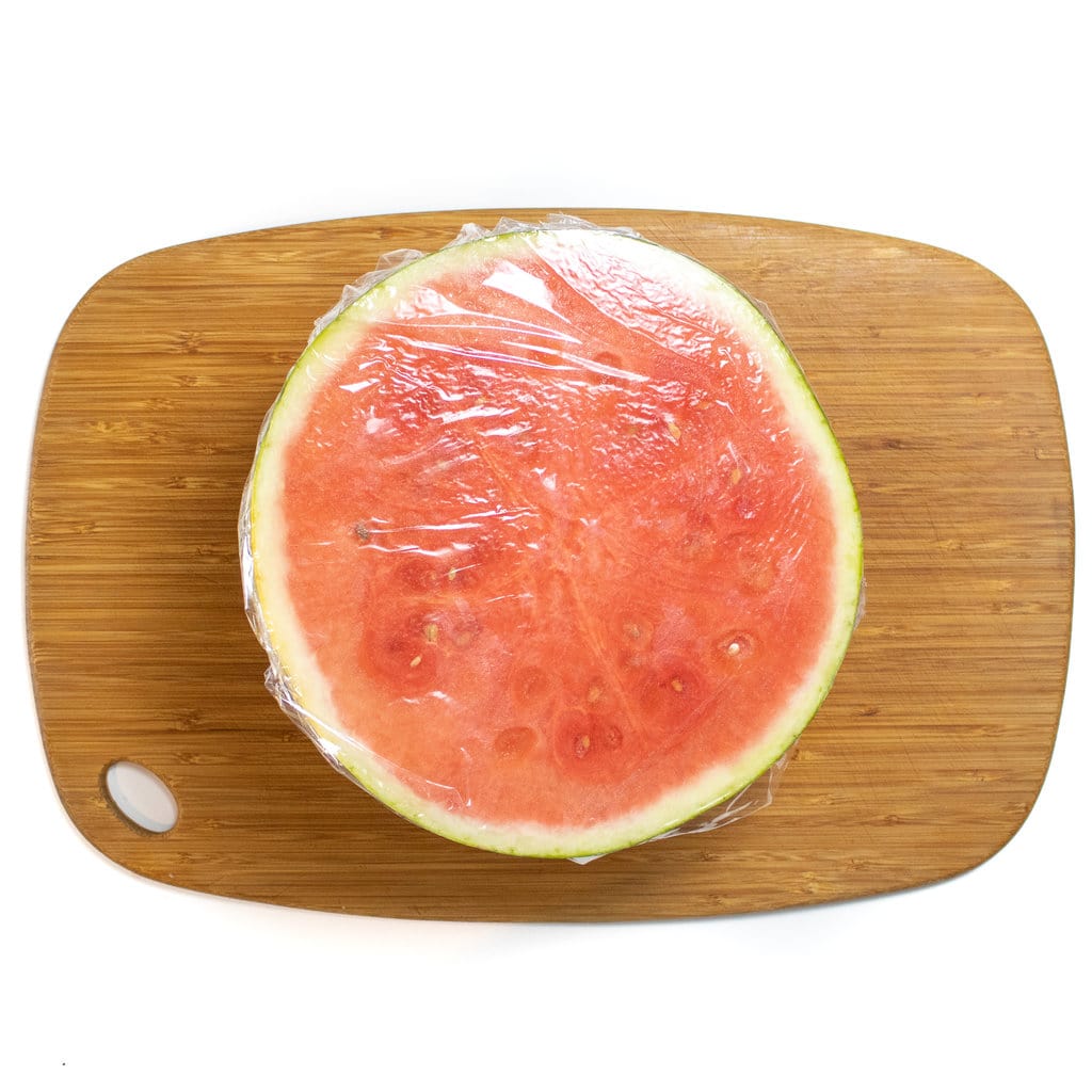 A half of a watermelon sitting on a cutting board with saran wrap on it.