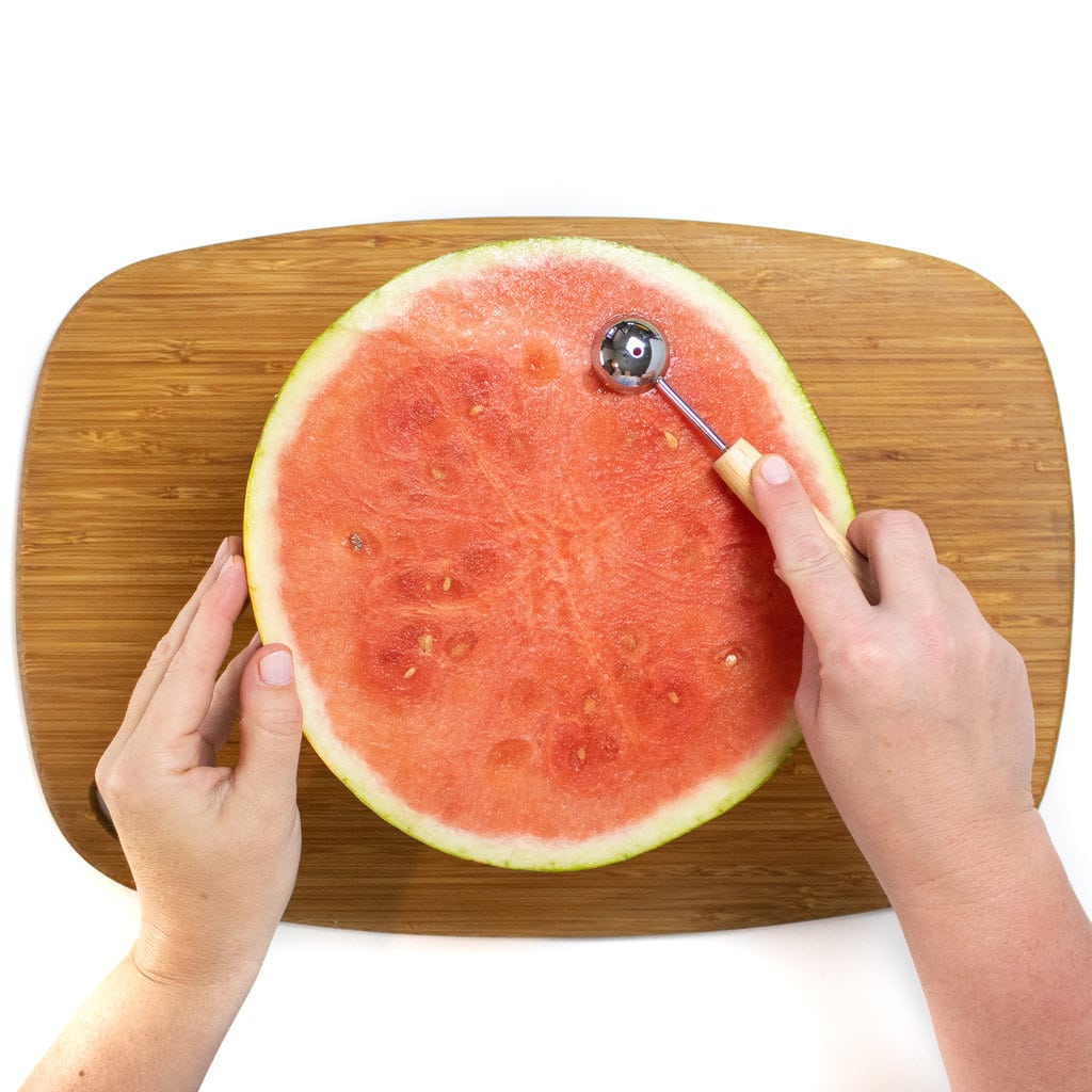 A watermelon cut in half with two hands holding a melon Baller over the flesh side of the watermelon.