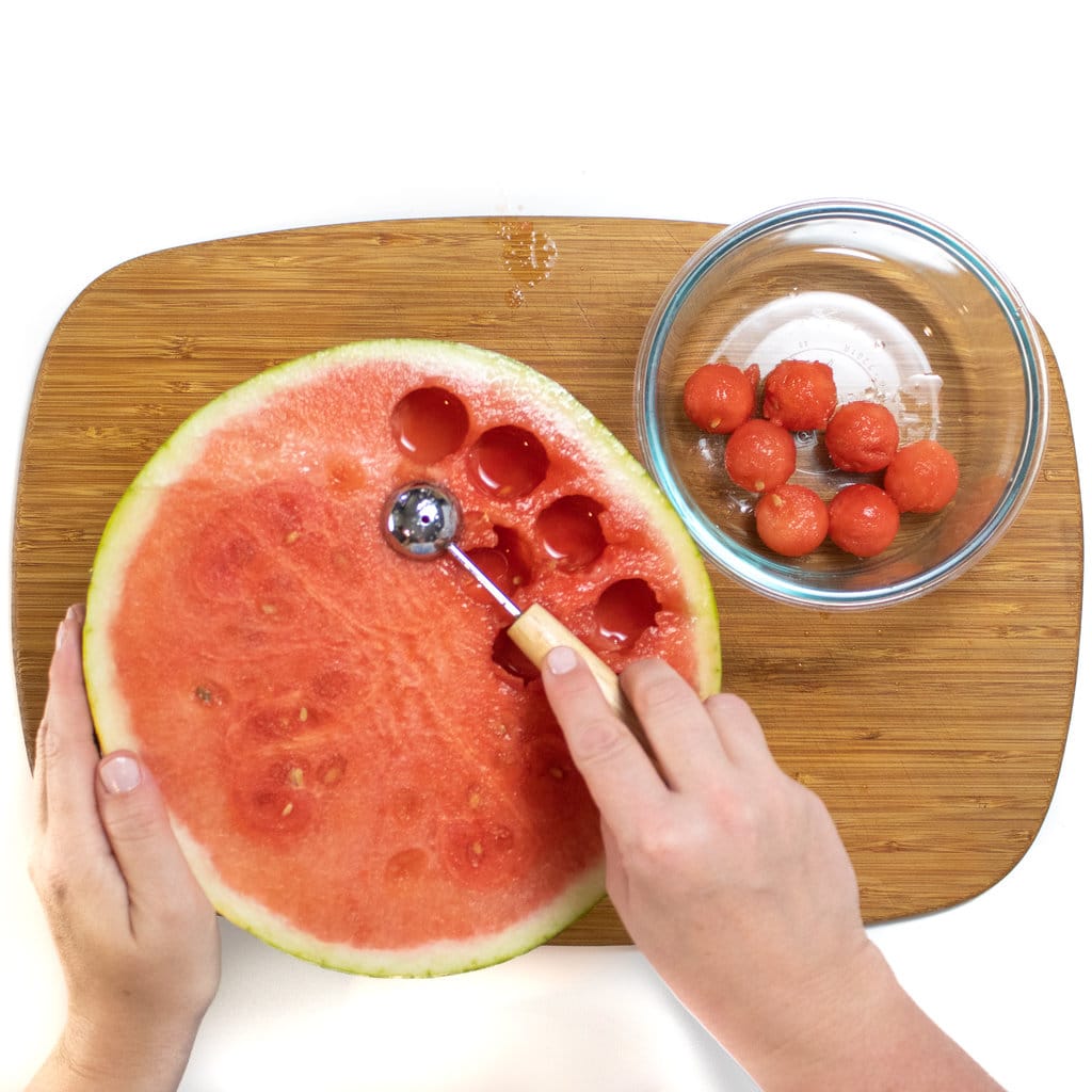 Half of a watermelon sitting on a cutting board with hands using a melon Baller with a bowl of melon balls next to it