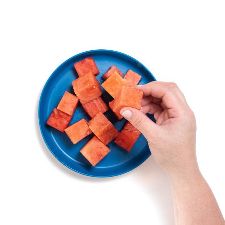 Blue plate with cubes of watermelon and a hand holding a cube of water melon.