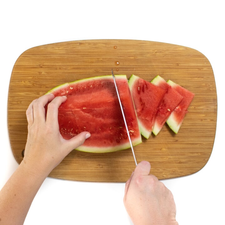 A wedge of a watermelon on a wooden cutting board with two hands, one hand holding a knife and cutting slices of watermelon.