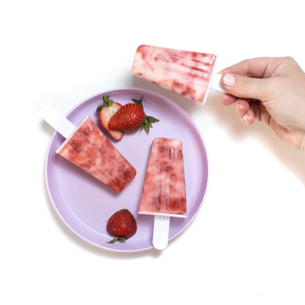 A kids purple plate with two strawberry yogurt popsicles and sliced strawberries on it and a hand holding another strawberry yogurt popsicle against a white background.