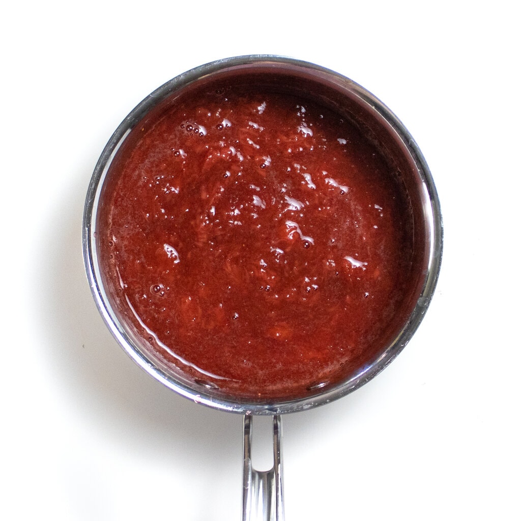Silver sauce pan on a white background filled with a simmered strawberry, maple syrup and vanilla pu