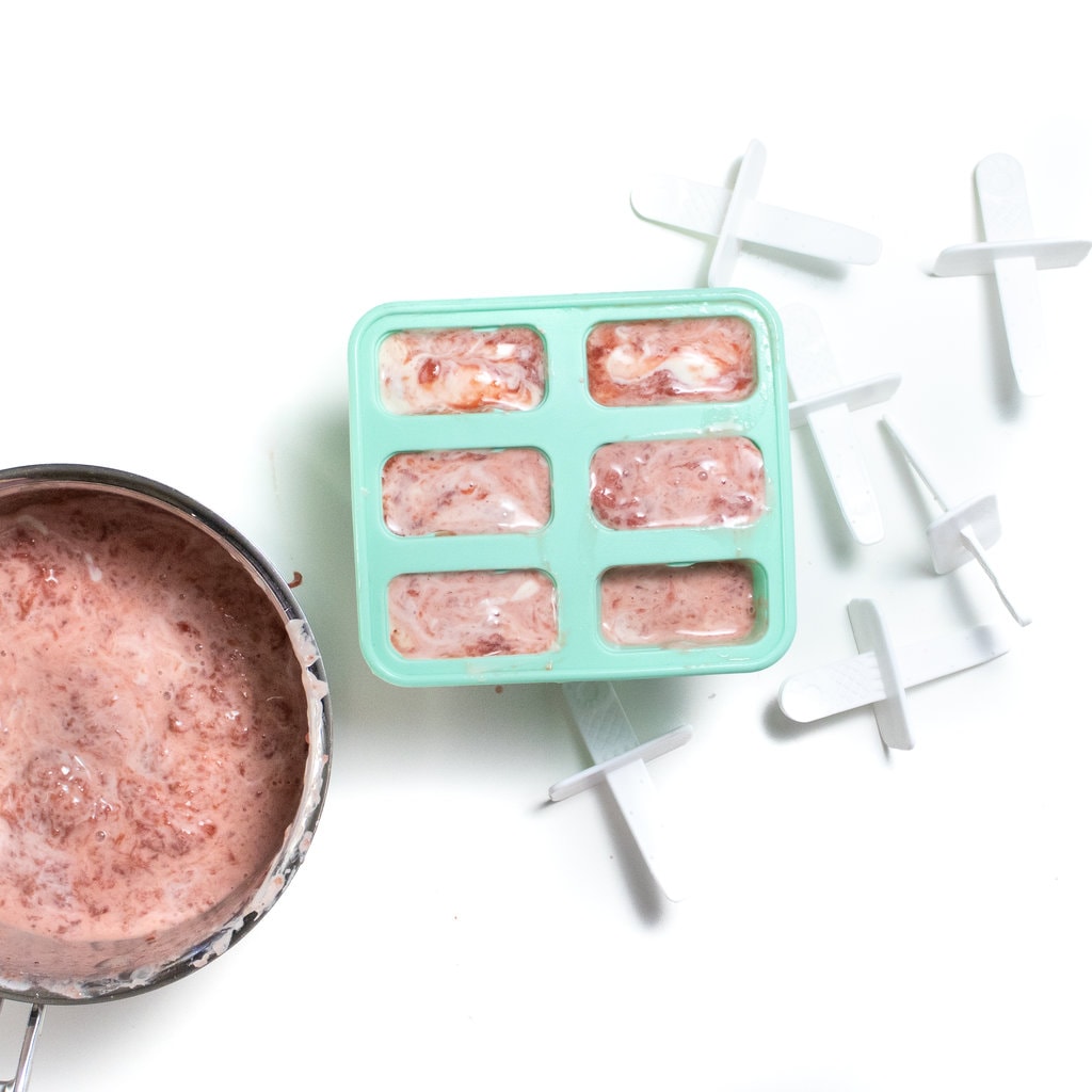 Spread of a pan filled with strawberry and yogurt and a popsicle mold with popsicle sticks on that white background.