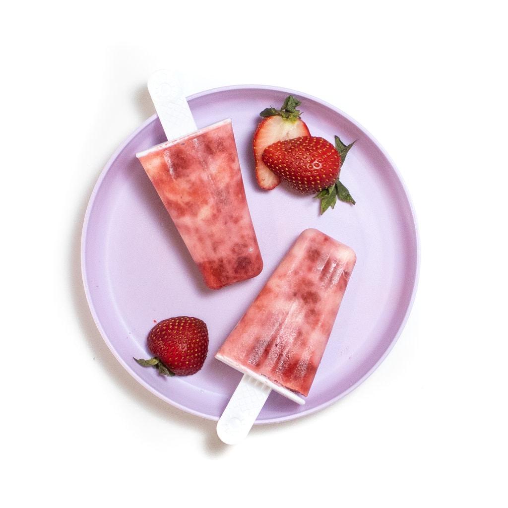 A purple kids plate with two strawberry yogurt popsicles and sliced strawberries on it against a white background.