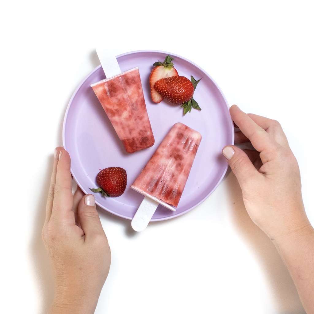 A purple kids plate with two hands holding it into strawberry yogurt popsicles and sliced strawberries.