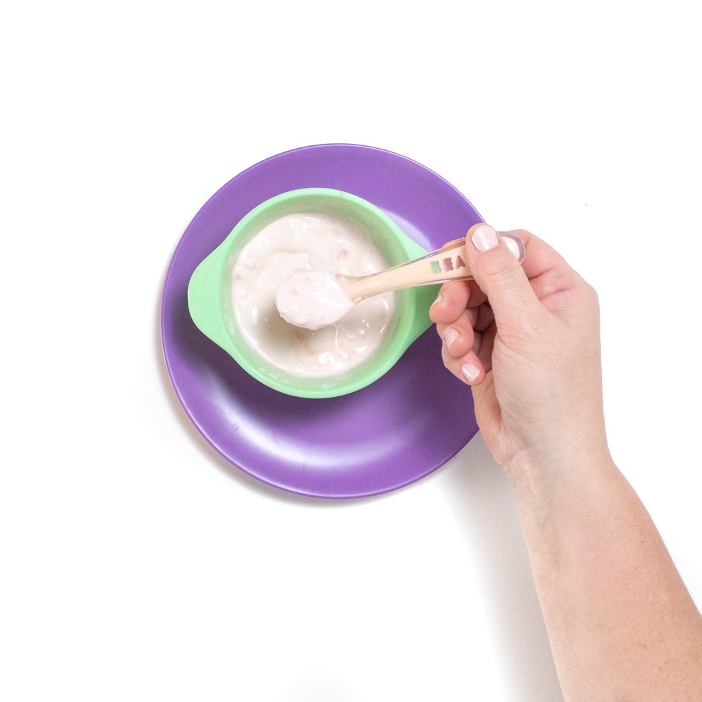 A hand holding a baby spoon with chunky watermelon purée and yogurt over a teal baby bowl and purple baby plate.