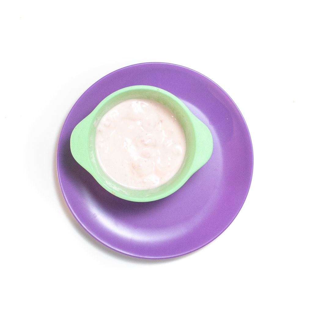 A teal baby bowl with chunky watermelon and yogurt purée on top of a purple plate against a white background.