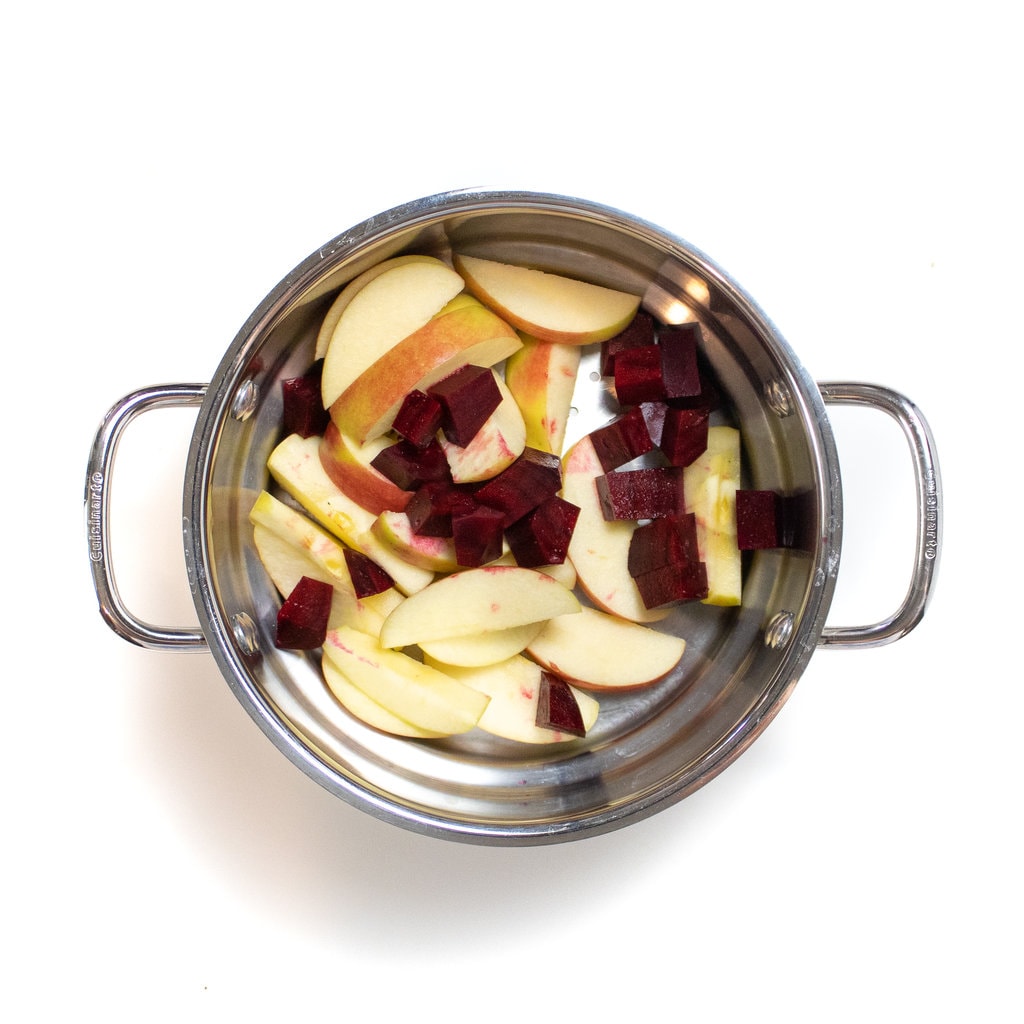 A steamer basket sitting on the white background filled with apples that have been sliced in chunks of red beets.