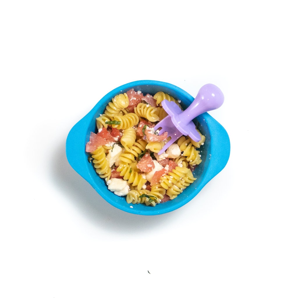 A blue baby bowl against a white background, the bowl is filled with pasta, watermelon, feta and basil with a purple baby fork resting inside.