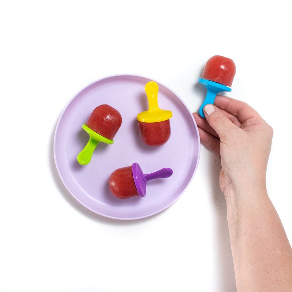 A purple kids plate with watermelon popsicles and a hand holding a blue popsicle stick with watermelon pop.