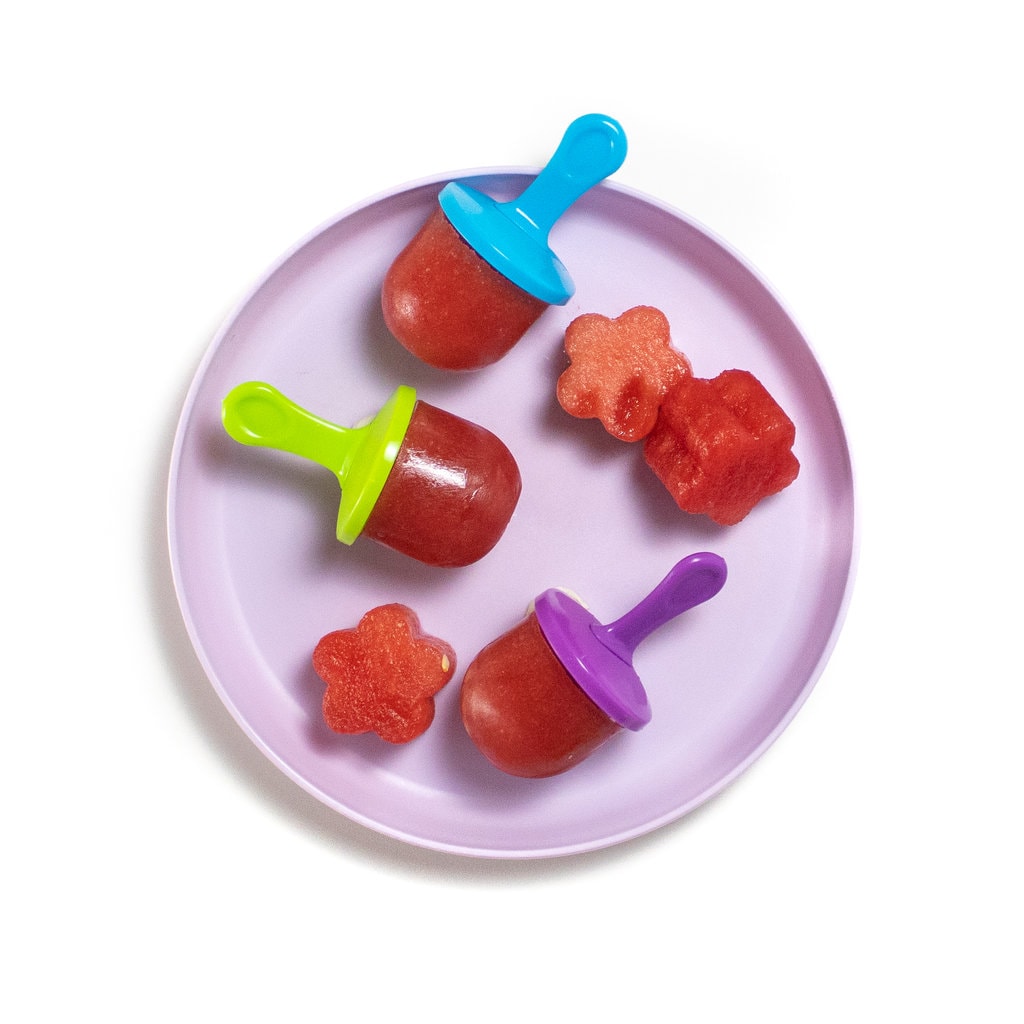 A purple kids plate on a white background with purple, green, blue popsicle sticks with watermelon popsicles and chunks of watermelon on the plate.