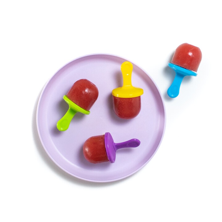 A purple kids plate on a white background with watermelon popsicles with colorful popsicle sticks.