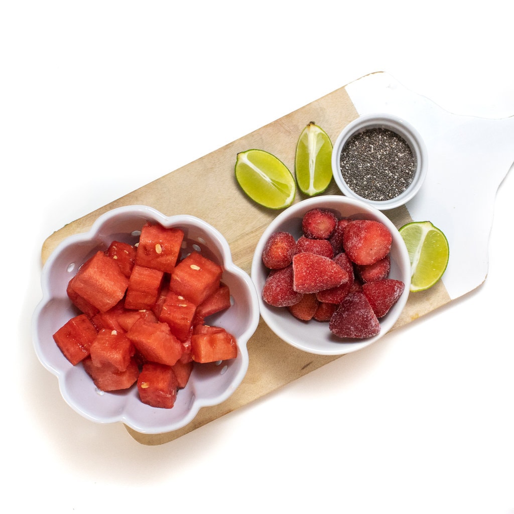 A wooden cutting board with a bowl of watermelon, a bowl of frozen strawberries, a bowl of Chia seeds and a cut up line.