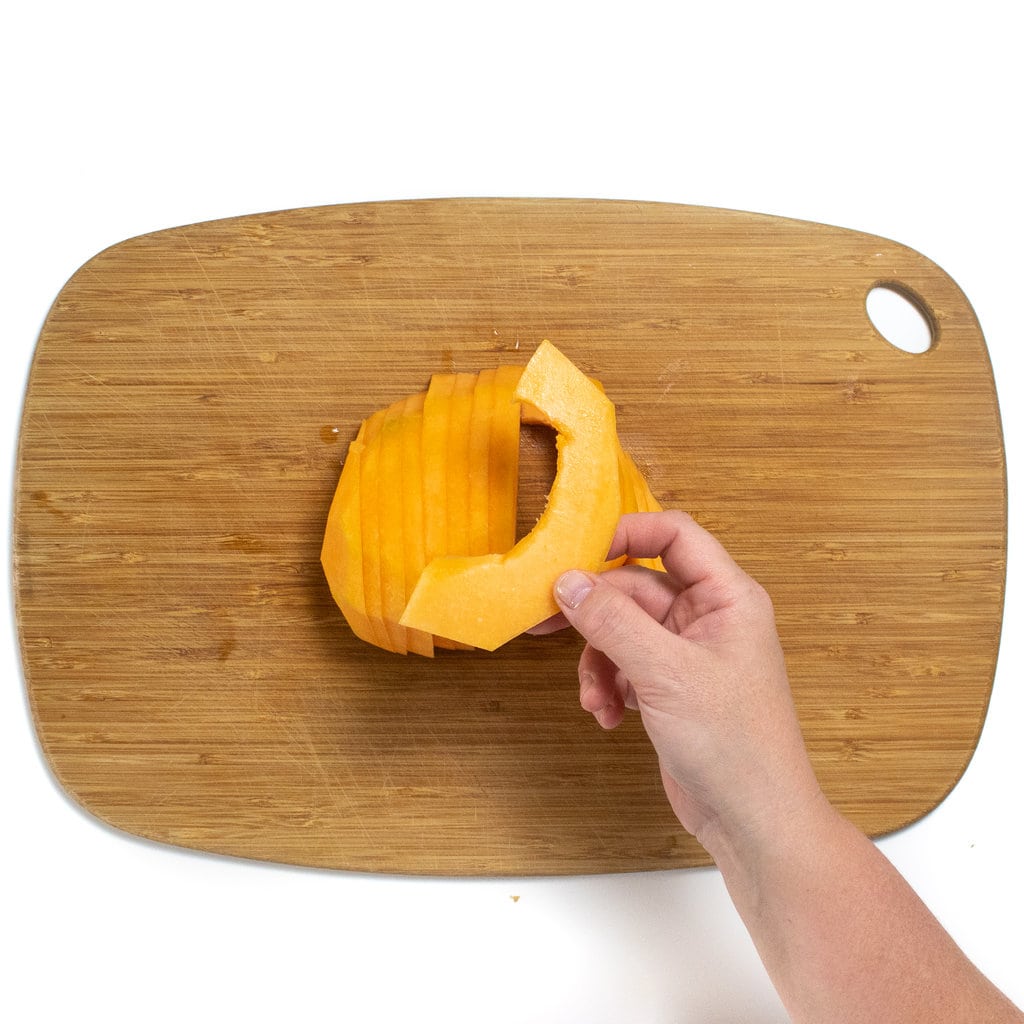 A hand holding a thin slice of sliced cantaloupe above a wooden cutting board against a white background.