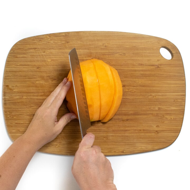 Hands holding a cantaloupe cutting it into thick wedges on a wooden cutting board against a white backdrop.