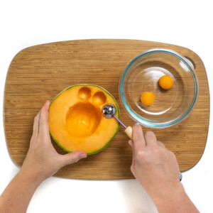 Two hands holding a Cantelope and using a scooper to form Cantaloupe balls.