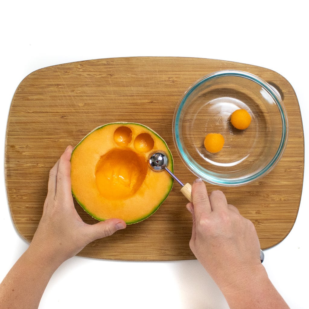 Two hands holding a Cantelope and using a scooper to form Cantaloupe balls.