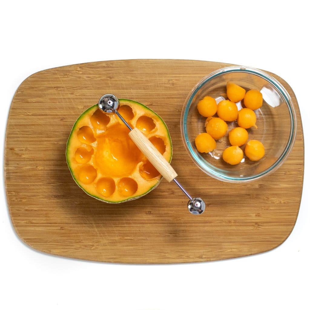 A wooden cutting board with a cantaloupe with scoops taken out and a glass container with cantaloupe balls.