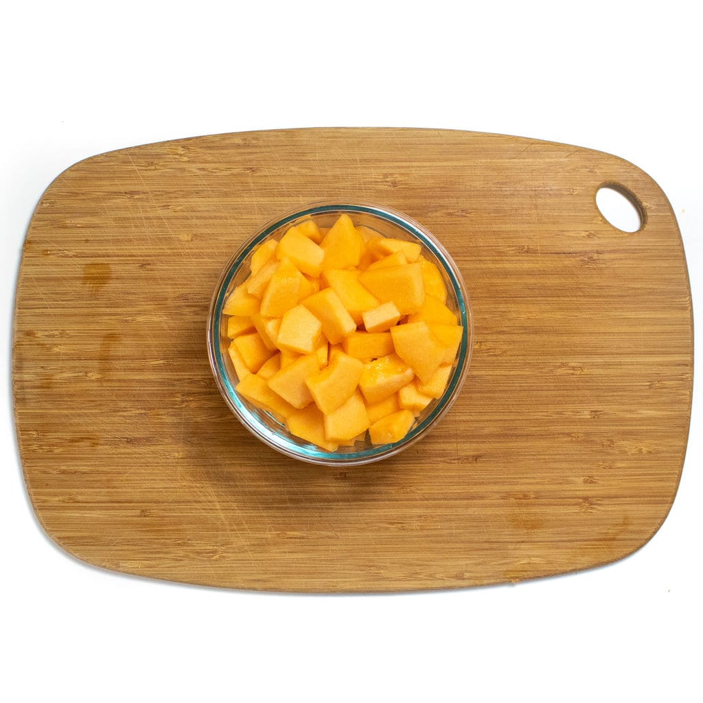 A wooden cutting board with a glass container with diced cantaloupe.