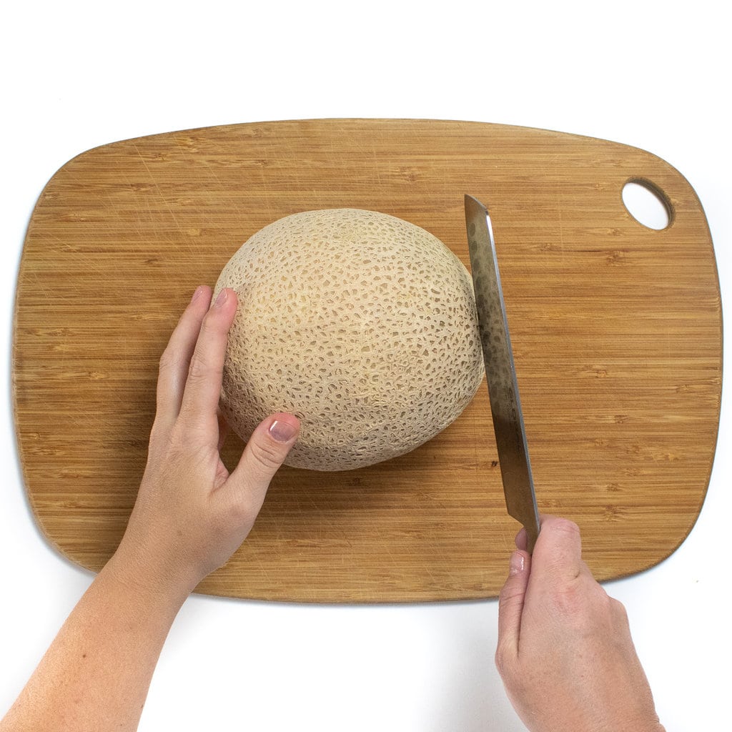 Wooden cutting board and a whole cantaloupe with two hands cutting the end off of one side.