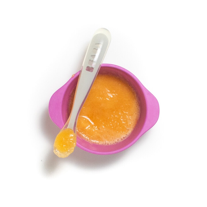 A pink baby bowl with puréed cantaloupe with a spoon resting on top with purée inside.