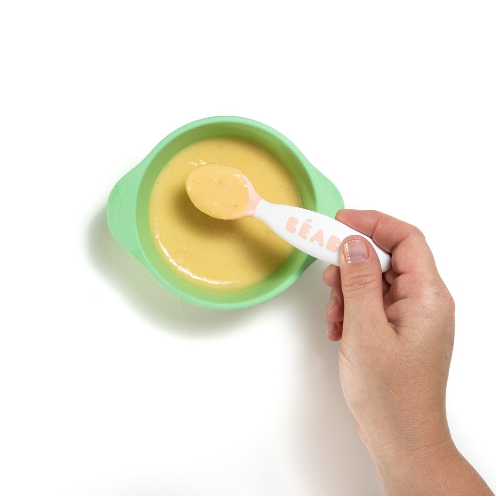 A teal baby bowl with a hand holding a baby spoon stirring a cantaloupe, yogurt and banana purée.