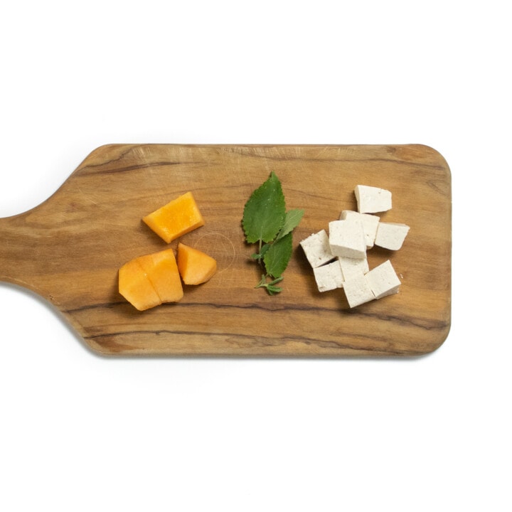 It's small wooden cutting board with chunks of cantaloupe, two leaves of mint and cubes of tofu.