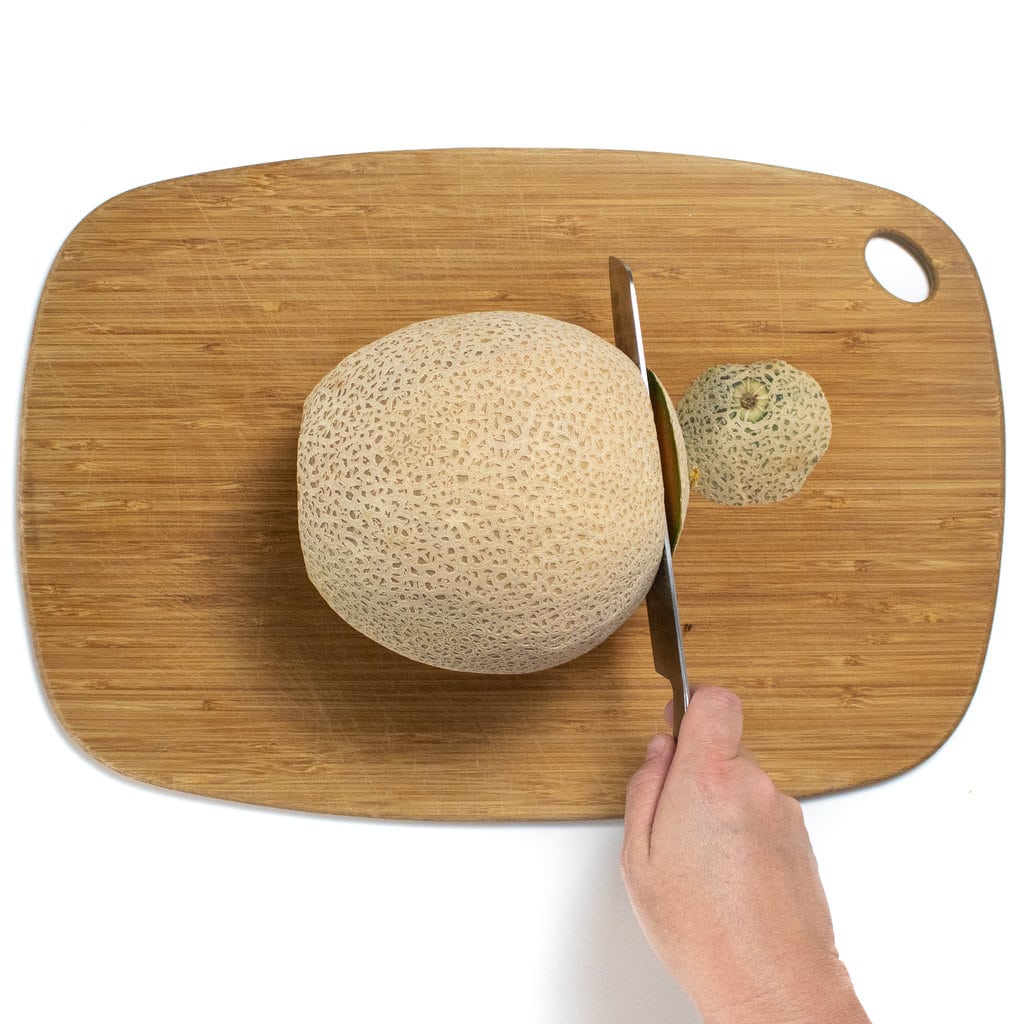 Cantaloupe melon on a cutting board with a knife cutting off one end of it.