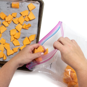 A baking sheet with frozen cantaloupe in hand putting the frozen cantaloupe into a Ziploc bag.