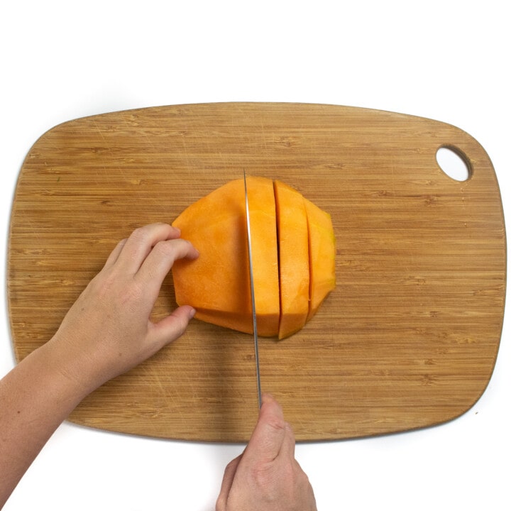 A wooden cutting board with hands cutting thick slices of cantaloupe.