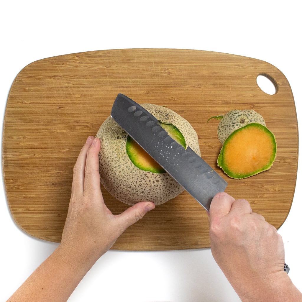 Two hands holding a cantaloupe in a knife cutting off the skin around the flesh of a cantaloupe on a cutting board.
