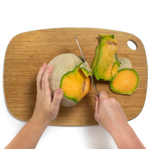 A pair of hands holding a cantaloupe in a knife cutting off the skin and exposing the orange flesh.