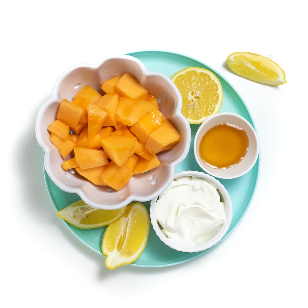 A teal blue kids plate with cantaloupe, yogurt, lemons and sweetener as a spread to show the ingradients for cantaloupe popsicles.
