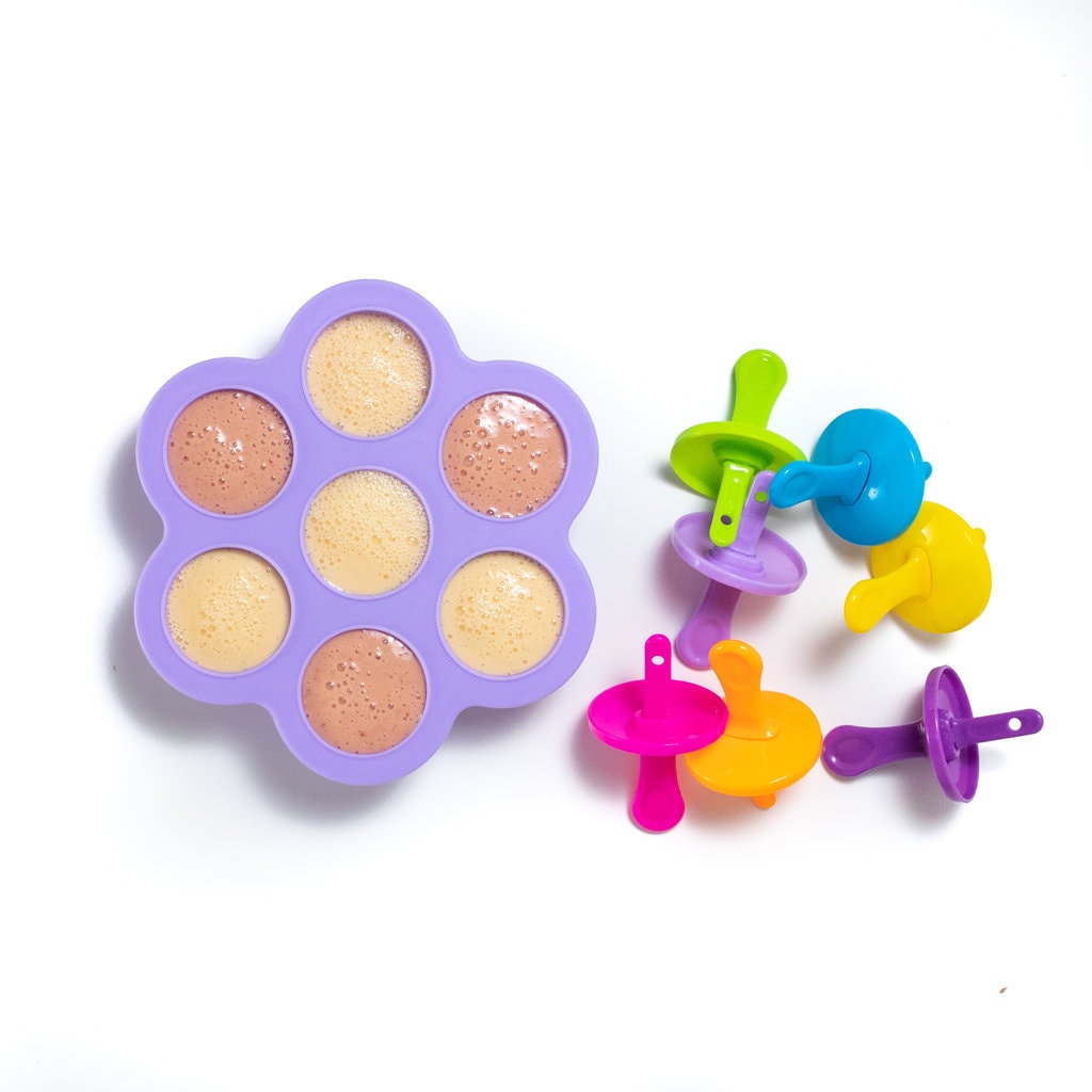 A purple popsicle tray with cantaloupe and strawberry popsicles for kids with colorful popsicle sticks on the side against a white background.