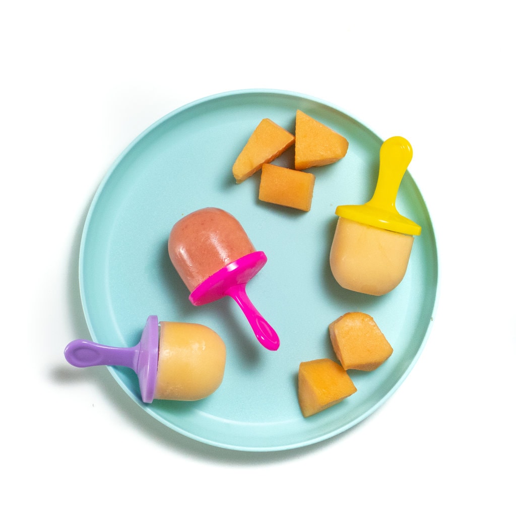 a teal kids play with colorful popsicle sticks and Cantelope Popsicles with the second option of strawberry cantaloupe Popsicles with chunks of cantaloupe on the plate and white background.