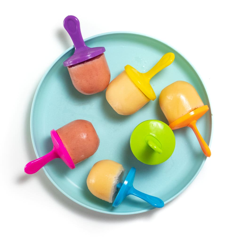 A teal kids played against a white background with colorful popsicle sticks and cantaloupe and strawberries cantaloupe popsicles scattered about.
