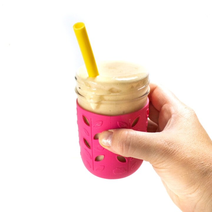 A hand holding a pink smoothie cup with cantaloupe smoothie inside and a yellow straw against the way background.