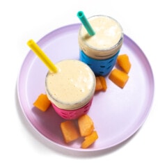 A purple plate with 2 cups and Cantelope scattered about the cups full of cantaloupe smoothie.