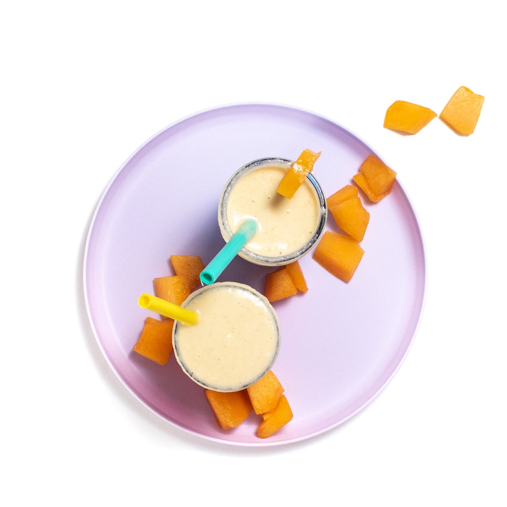 A purple plate with 2 cups and Cantelope scattered about the cups full of cantaloupe smoothie.