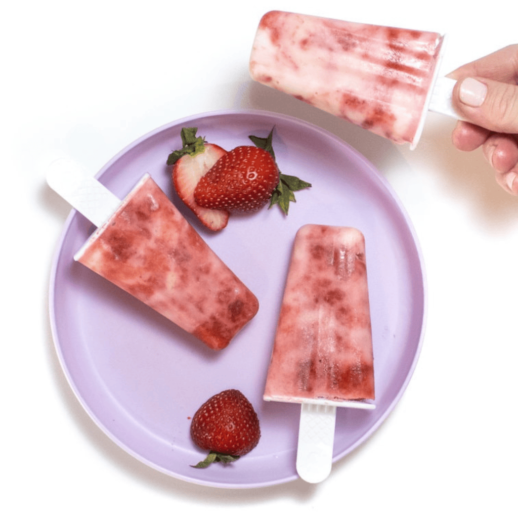 Hand holding a strawberry yogurt popsicle over a white counter and a purple kids plate full of more popsicles and sliced strawberries.