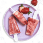 purple kids plate on a marble kitchen counter with three strawberry popsicles with swirled greek yogurt on the plate with sliced strawberries.