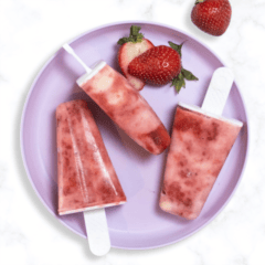 purple kids plate on a marble kitchen counter with three strawberry popsicles with swirled greek yogurt on the plate with sliced strawberries.